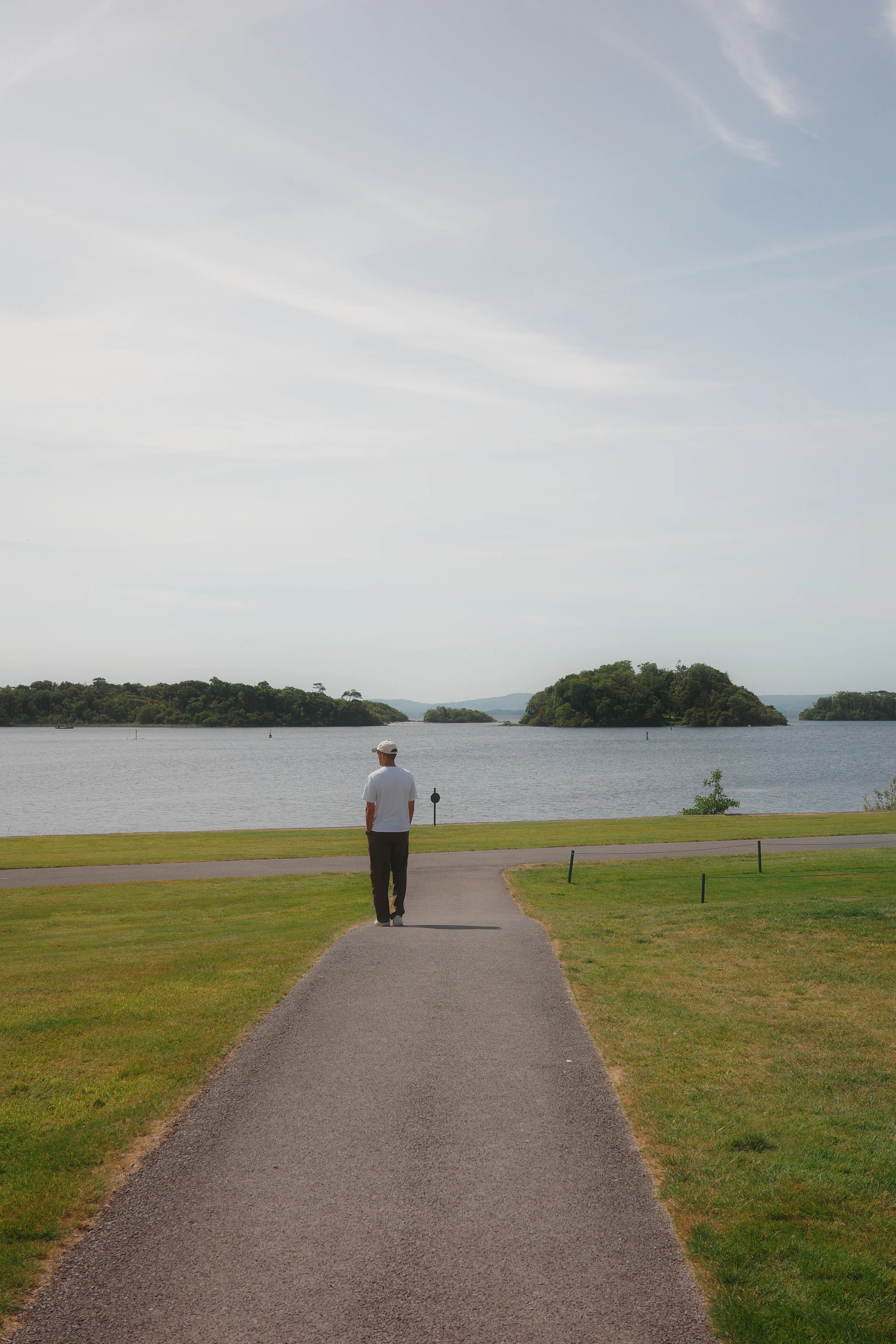 A man in a white shirt and hat walks down a paved path across a lawn toward a calm lake dotted with small wooded islands