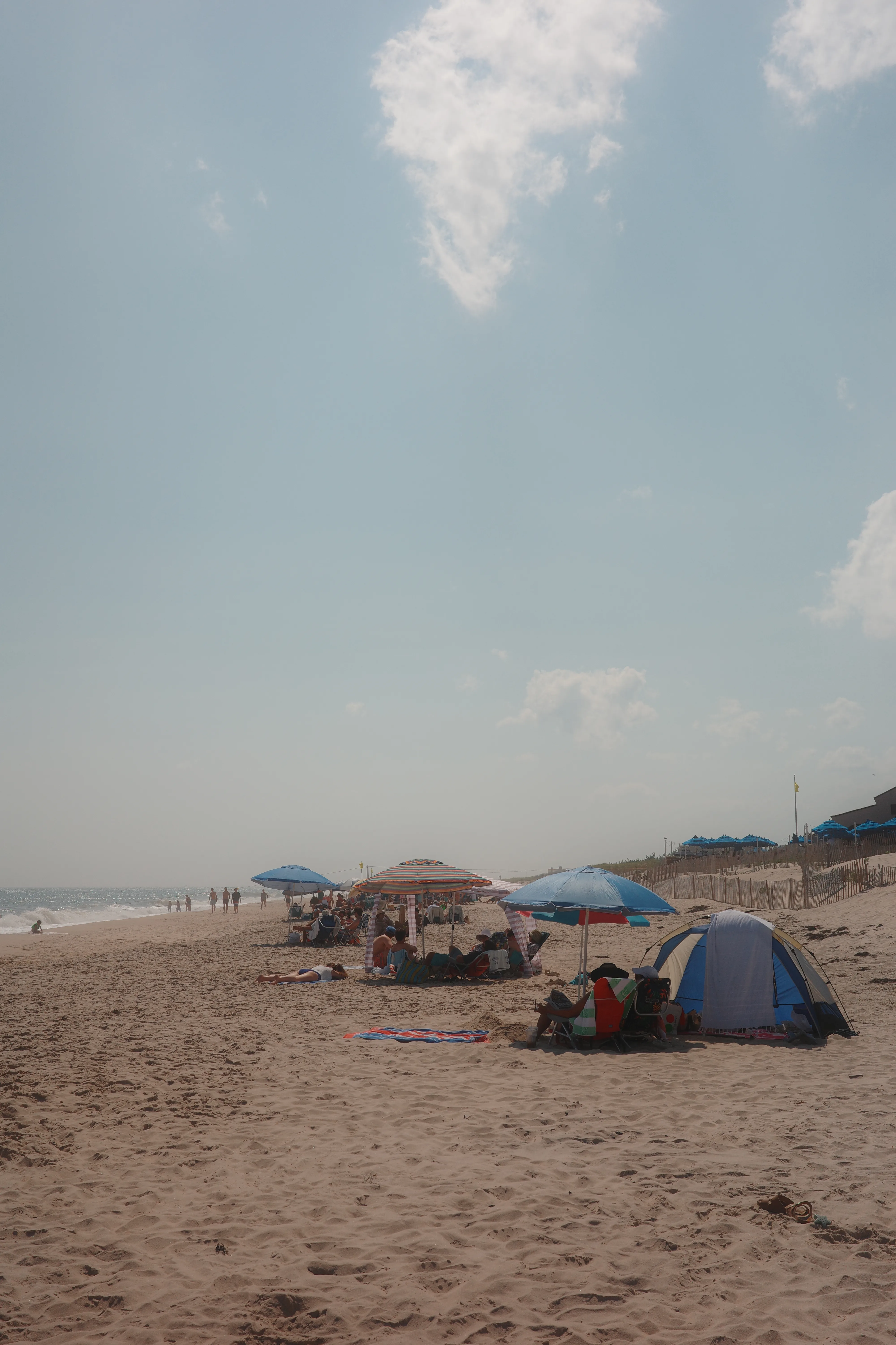 A hazy summer beach with blue umbrellas, a small tent, and beachgoers scattered along the shoreline under a pale sky