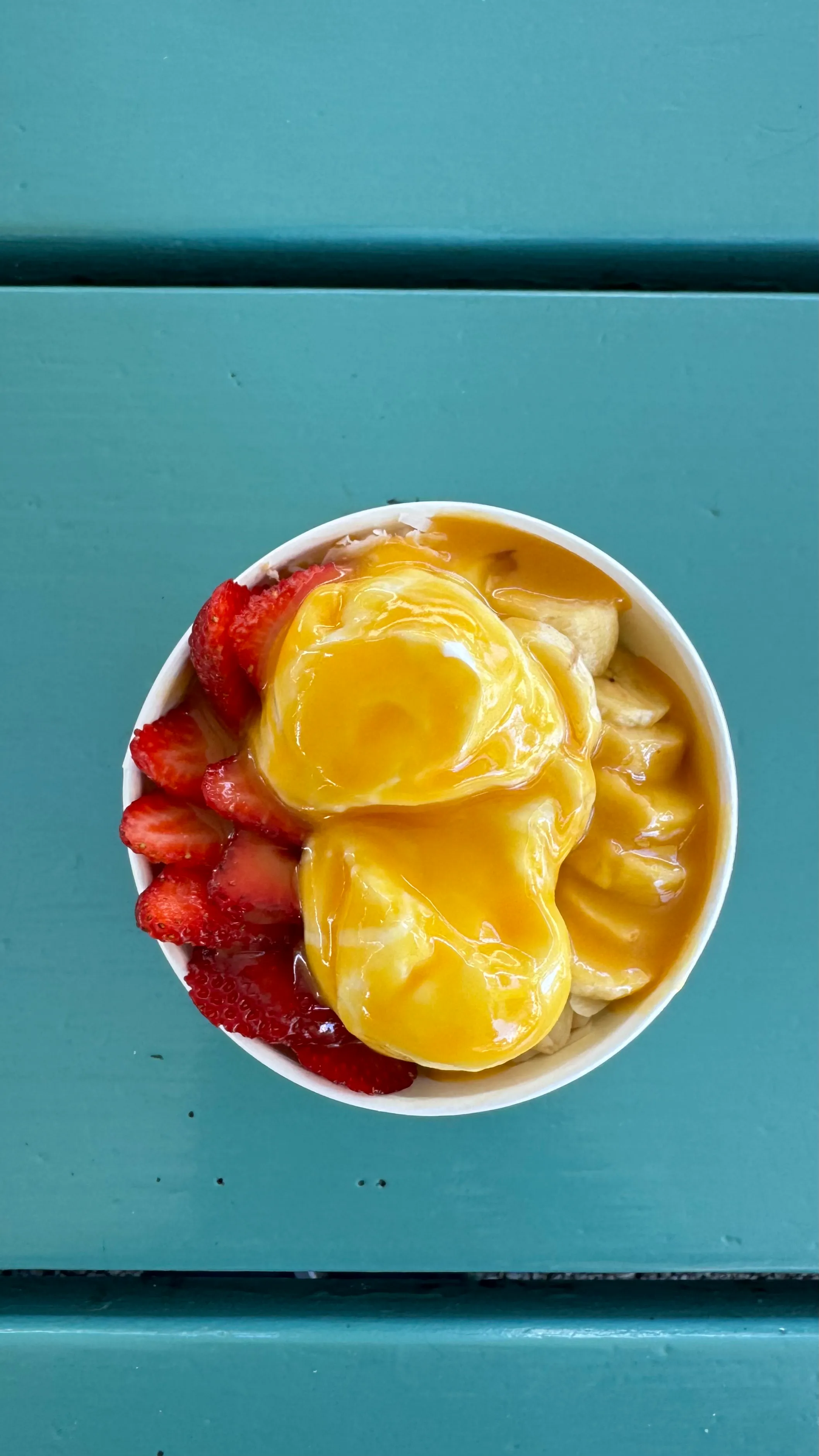 Overhead view of an açaí bowl topped with sliced strawberries, banana, and mango purée on a teal wooden table