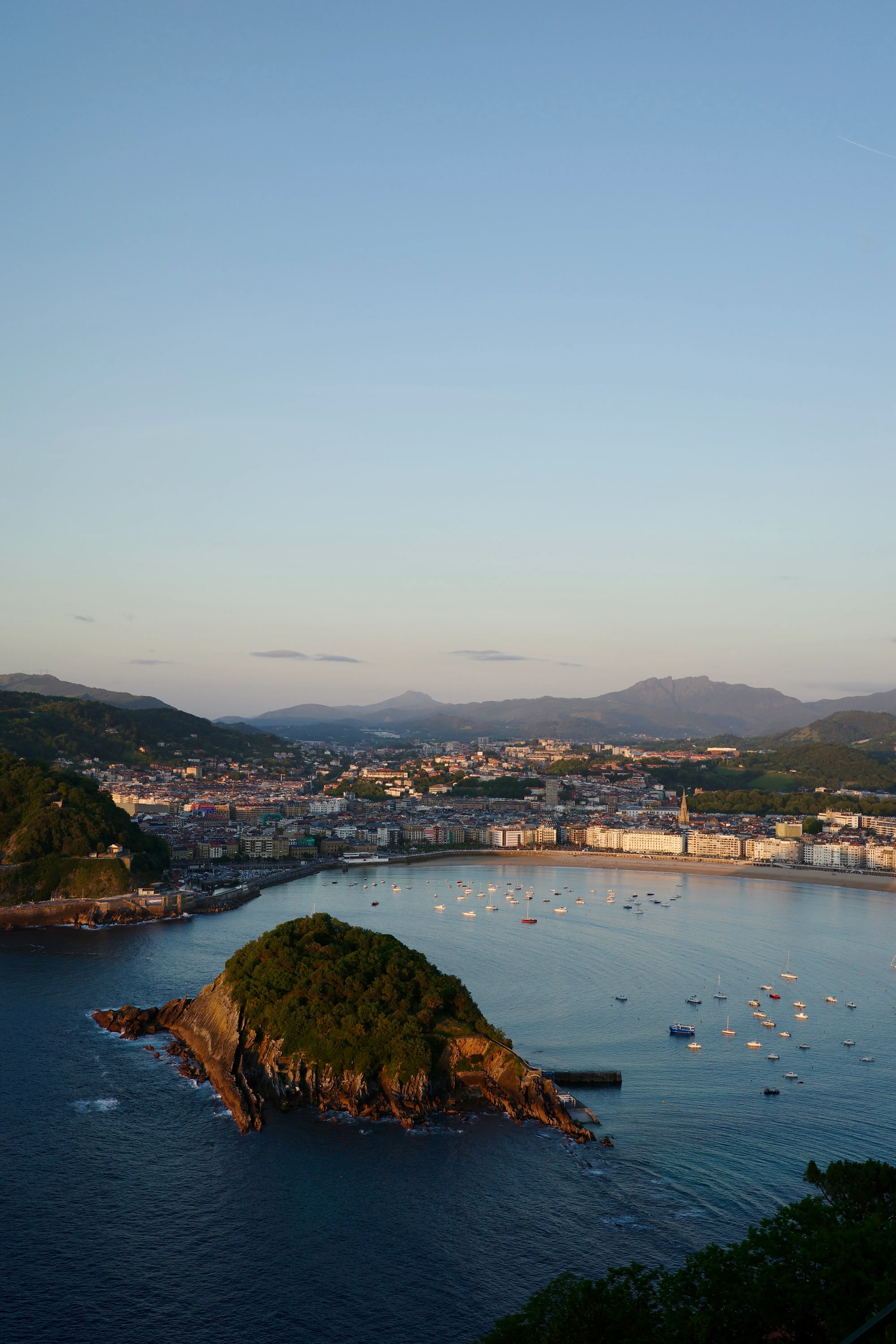 View from above of La Concha bay at dusk, with Santa Clara island in the foreground, sailboats on still water, and the city and mountains beyond