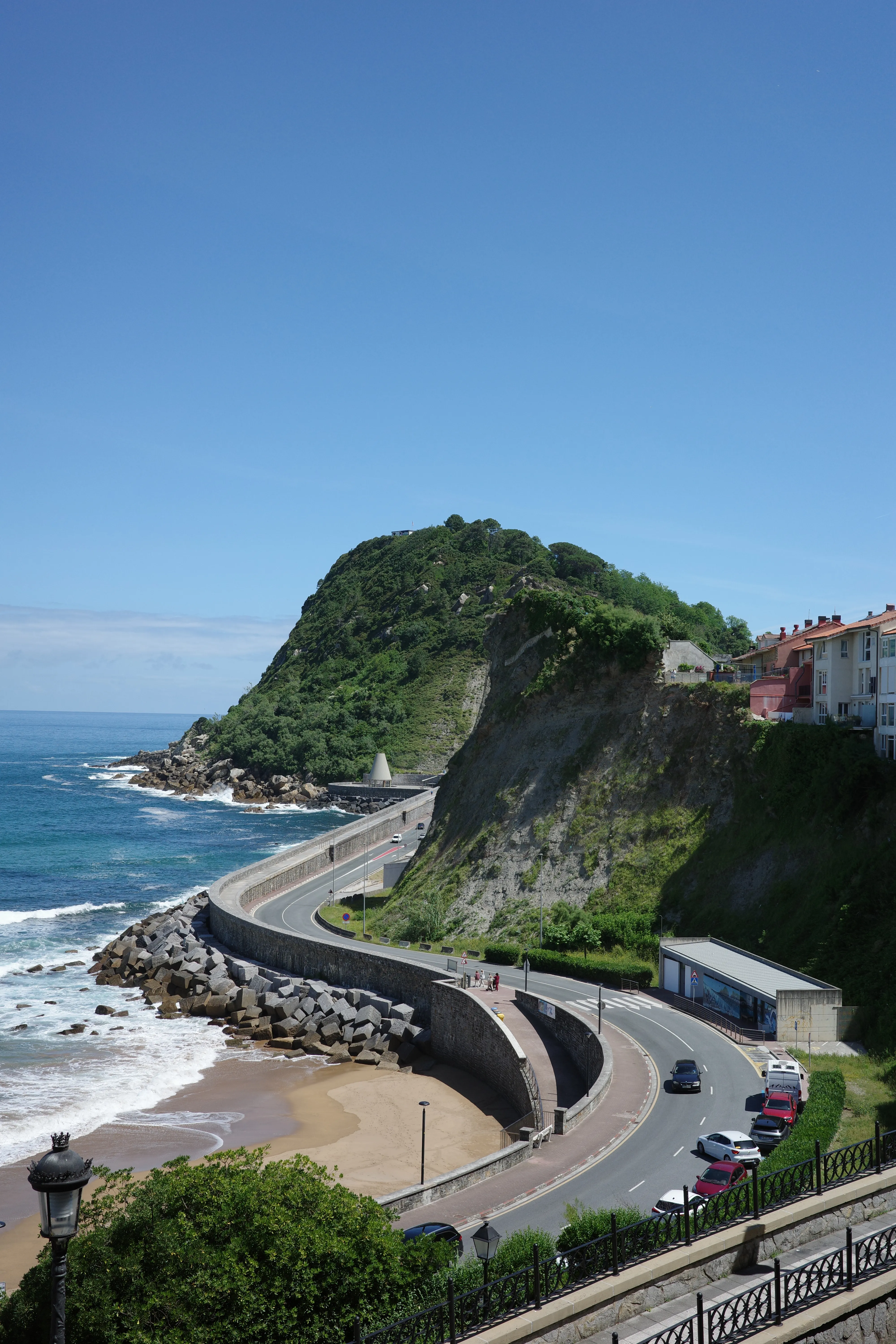 A coastal road curving along a seawall beneath a green headland, with waves breaking on rocks and a small beach below